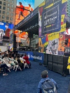 Audience facing an outdoor stage festooned with posters of Broadway shows.